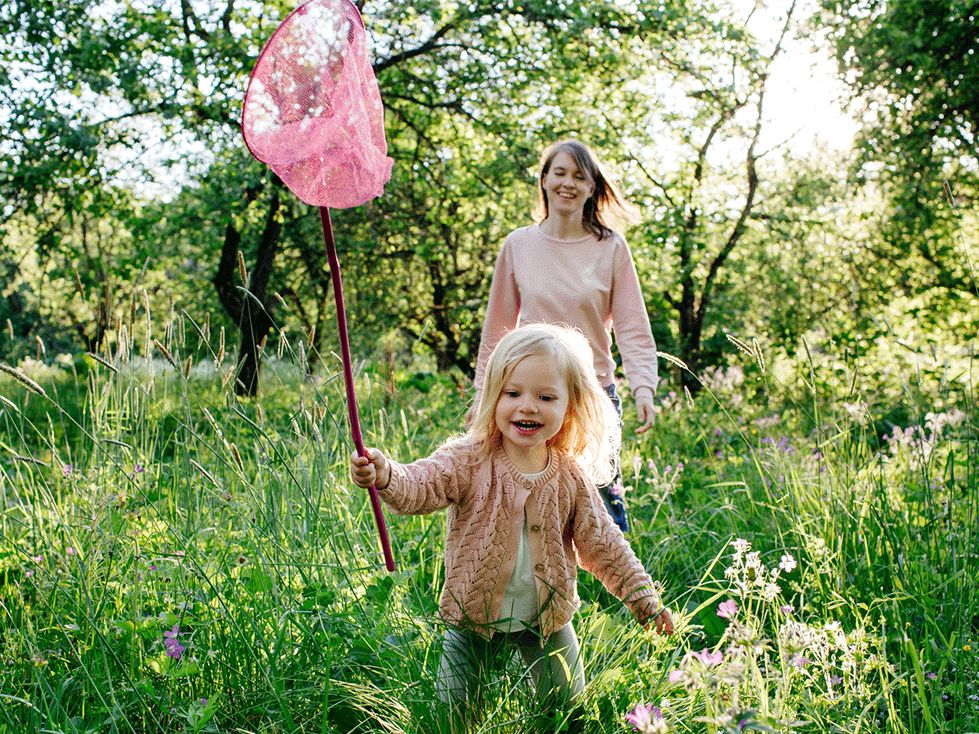 A little girl running through a meadow with a butterfly net, her mum smiling behind her